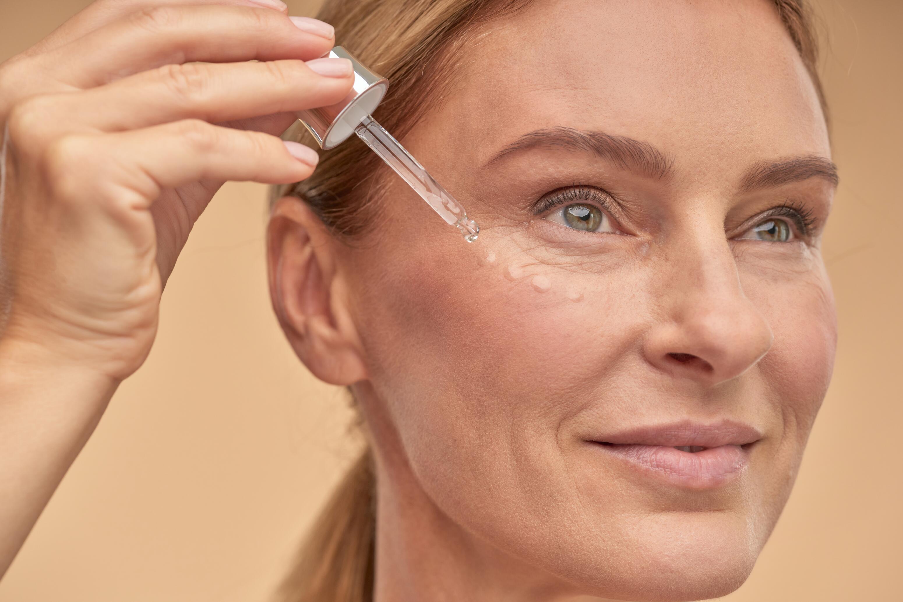 Nahaufnahme einer älteren, blonden Frau, sie hält eine Pipette und tropft eine Flüssigkeit unter ihre Augen.